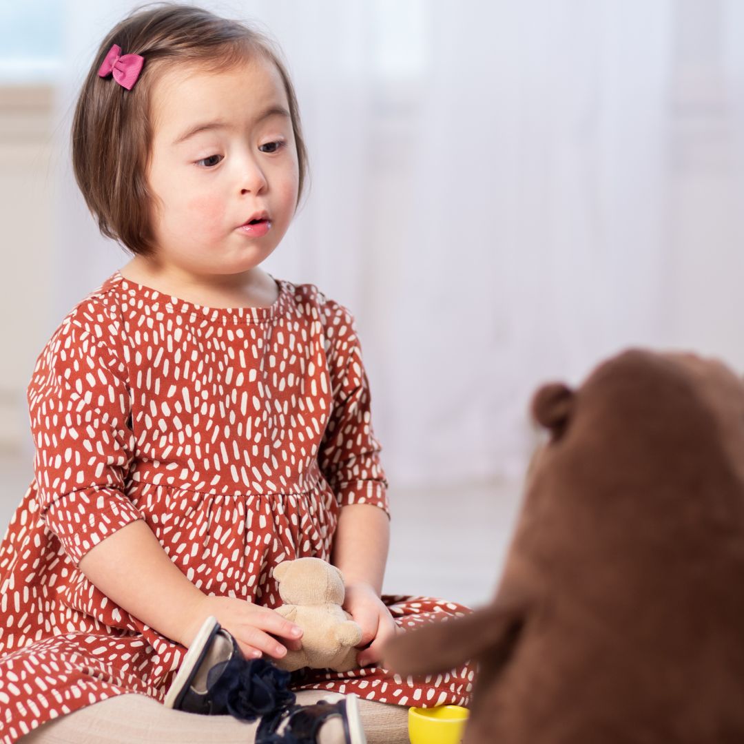 Das Bild zeigt ein kleines Mädchen mit kurzen braunen Haaren und einer rosa Haarspange. Sie trägt ein orangefarbenes Kleid mit weißen Mustern und hält ein Stofftier in der Hand.