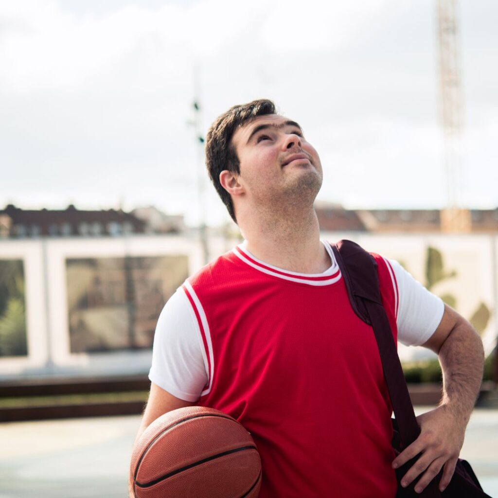 Das Bild zeigt einen jungen Mann im roten Basketballtrikot, der einen Basketball hält. Er hat dunkle Haare und schaut nach oben. Er trägt eine Sporttasche über der Schulter und steht im Freien vor einem unscharfen städtischen Hintergrund.