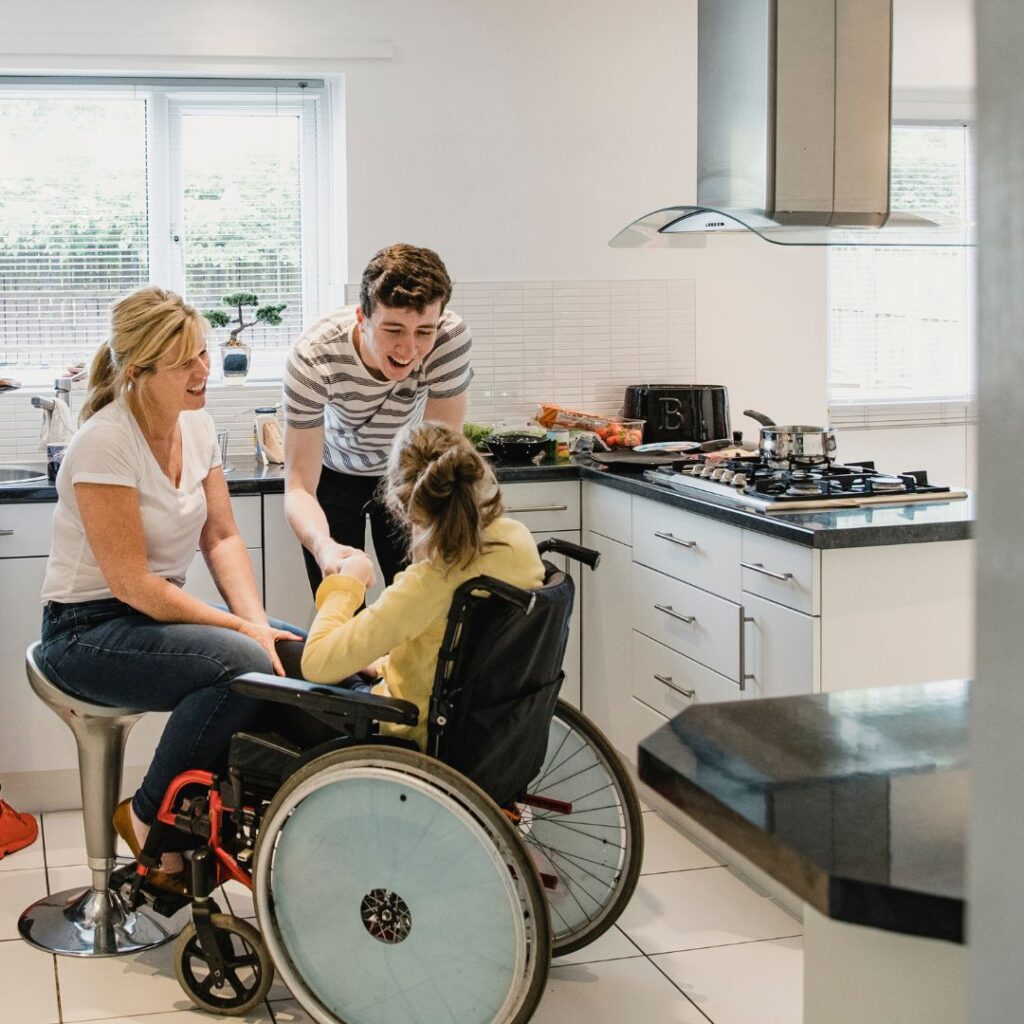 Das Bild zeigt eine fröhliche Szene in einer modernen Küche mit drei Personen: eine Frau auf einem Barhocker, einen jungen Mann und eine Person im Rollstuhl. Sie unterhalten sich und lächeln.